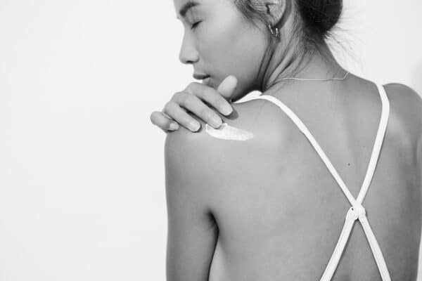 Black and white image of a woman gently applying skincare product to her shoulder slightly looking back.